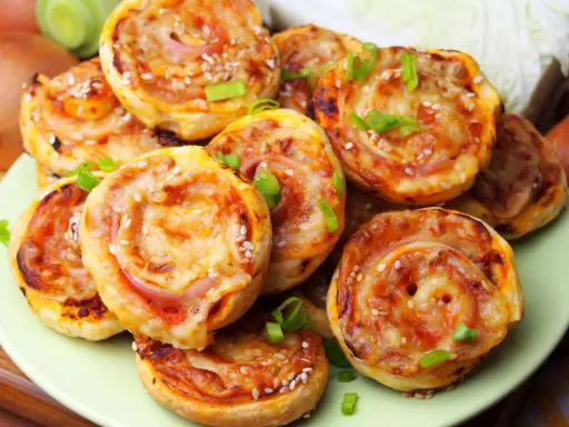 Pyszne pizzerinki z ciasta francuskiego, posypane sezamem i szczypiorkiem, ułożone na talerzu.