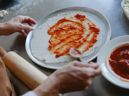 Ręce rozprowadzają pyszny sos do pizzy na cieście. Obok leży wałek i miseczka z sosem.