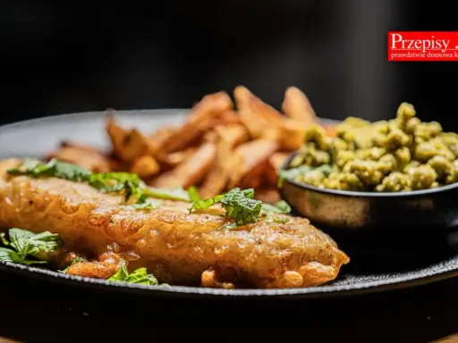 Chrupiący fish and chips z zielonym groszkiem i świeżą miętą na czarnym talerzu.