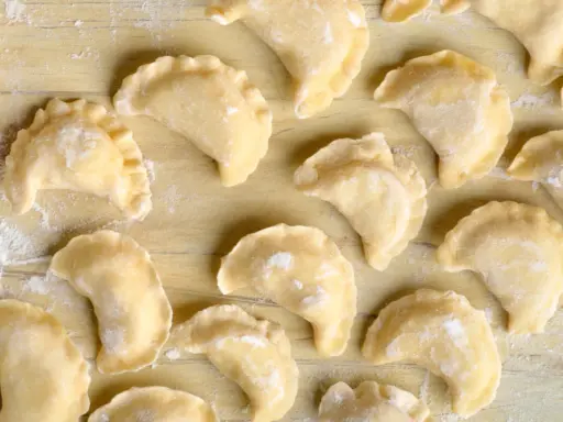 Surowe pierogi z farszem na pierogi ruskie, ułożone na drewnianym blacie, posypane mąką.