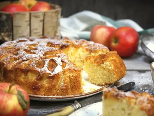 Pyszne ciasto z jabłkami, posypane cukrem pudrem, z jabłkami w koszyku w tle. Idealne na jesienny deser.