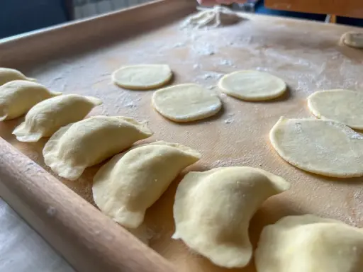 Gotowe do lepienia pierogi ruskie. Na drewnianej desce leżą okrągłe placki ciasta i uformowane pierożki.