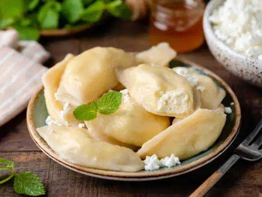 Pyszne pierogi z serem na słodko, posypane twarogiem i udekorowane miętą. Obok słoiczek miodu i miseczka z twarogiem.