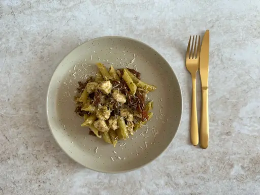 Penne z kurczakiem i suszonymi pomidorami, polane aromatycznym makaronem z pesto ze słoika, posypane parmezanem. Obok złote sztućce.