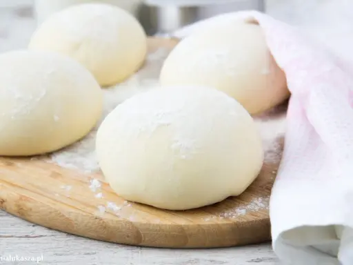 Kulki ciasta na spód do pizzy, posypane mąką, leżą na drewnianej desce. Obok leży różowy ręcznik.