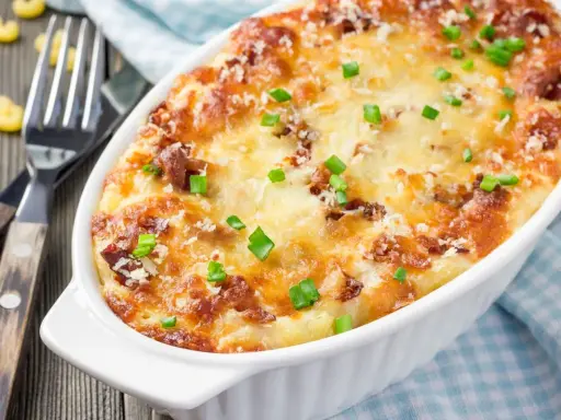 Pyszne, zapiekane mac and cheese w białym naczyniu, posypane szczypiorkiem i parmezanem. Obok widelce i luźne makarony.