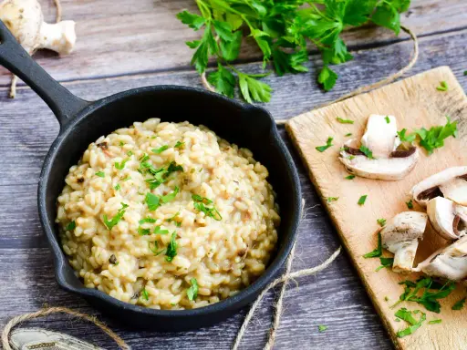 Kremowe risotto z warzywami, posypane natką pietruszki, obok świeże pieczarki na desce.