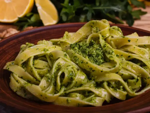 Tagliatelle z aromatycznym pesto z rukoli, podane na brązowym talerzu, z cytryną i orzechami w tle.