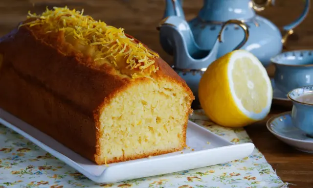 Puszysta babka piaskowa cytrynowa, ozdobiona skórką cytrynową, obok połówki cytryny i filiżanki herbaty.