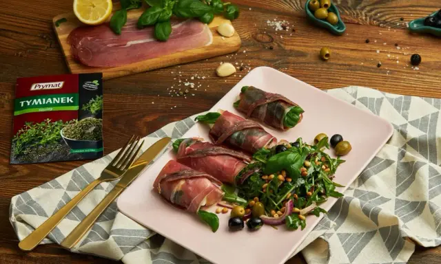 Roladki z szynką parmeńską i anchois, podane z rukolą, oliwkami i orzeszkami pinii. Obok talerza złote sztućce i opakowanie tymianku.