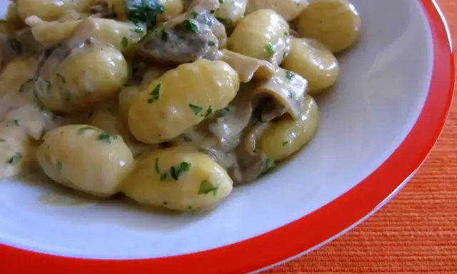 Pyszne gnocchi z sosem grzybowym i natką pietruszki, podane w białej misce z pomarańczowym rantem.