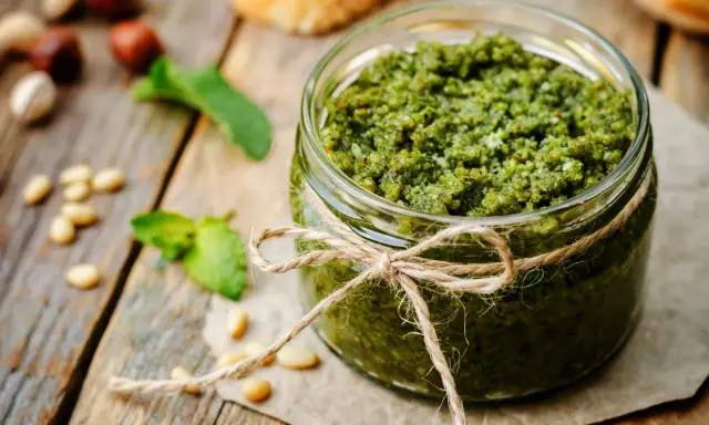 Słoik domowego pesto z bazylii, przewiązany sznurkiem. Obok orzeszki piniowe i listki bazylii.