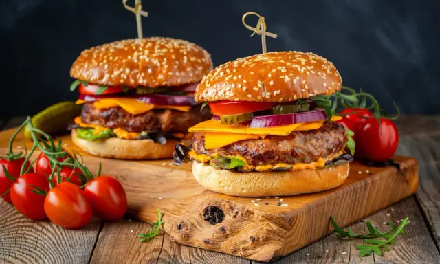 Dwa soczyste burgery z idealnie wysmażonym mięsem na burgera, serem, warzywami i sezamową bułką, podane na drewnianej desce z pomidorkami koktajlowymi.