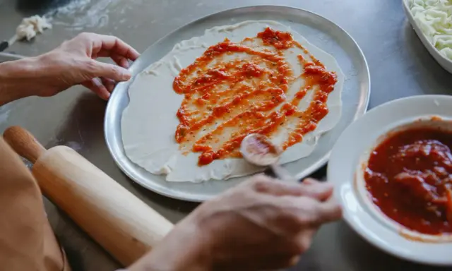 Ręce rozprowadzają pyszny sos do pizzy na cieście. Obok leży wałek i miseczka z sosem.