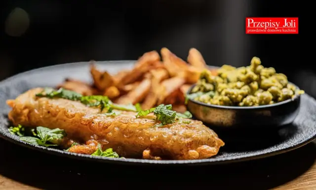 Chrupiący fish and chips z zielonym groszkiem i świeżą miętą na czarnym talerzu.
