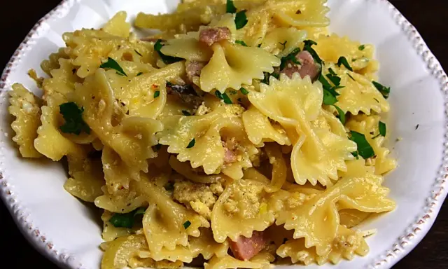 Pyszny makaron farfalle z jajkiem, kawałkami szynki i posiekaną natką pietruszki, podany w białej misce.