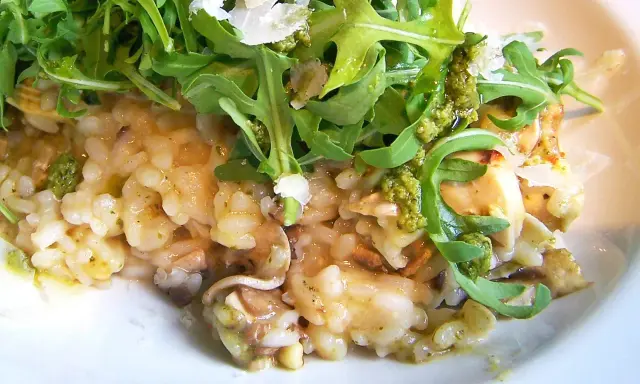 Kremowe risotto z pieczarkami, polane zielonym pesto i posypane parmezanem, udekorowane świeżą rukolą.