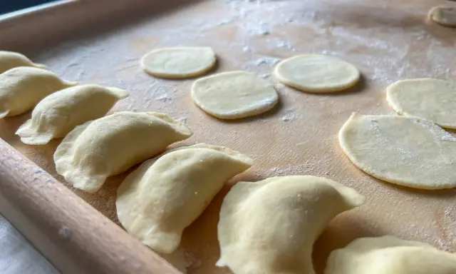 Gotowe do lepienia pierogi ruskie. Na drewnianej desce leżą okrągłe placki ciasta i uformowane pierożki.