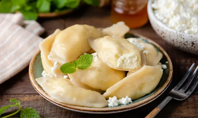 Pyszne pierogi z serem na słodko, posypane twarogiem i udekorowane miętą. Obok słoiczek miodu i miseczka z twarogiem.
