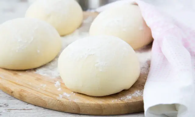 Kulki ciasta na spód do pizzy, posypane mąką, leżą na drewnianej desce. Obok leży różowy ręcznik.