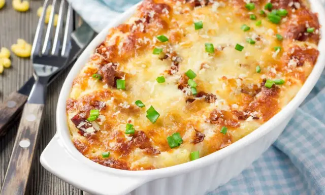 Pyszne, zapiekane mac and cheese w białym naczyniu, posypane szczypiorkiem i parmezanem. Obok widelce i luźne makarony.