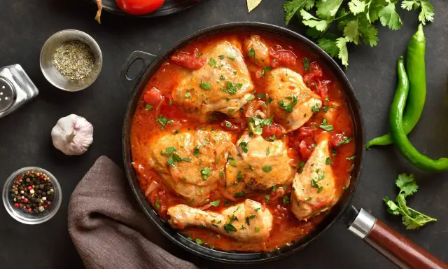 Pyszny kurczak w sosie pomidorowym, duszony z cebulą i przyprawami, udekorowany natką pietruszki. Obok zielona papryczka chili i pomidory.