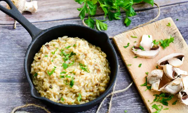 Kremowe risotto z warzywami, posypane natką pietruszki, obok świeże pieczarki na desce.