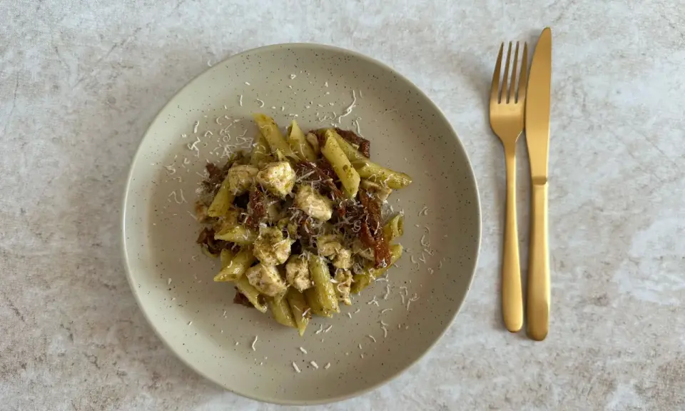 Penne z kurczakiem i suszonymi pomidorami, polane aromatycznym makaronem z pesto ze słoika, posypane parmezanem. Obok złote sztućce.