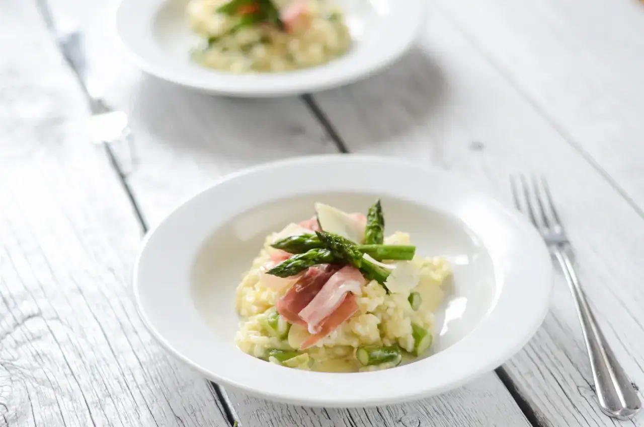 Pyszne risotto ze szparagami, udekorowane szynką parmeńską i parmezanem, podane na białym talerzu.