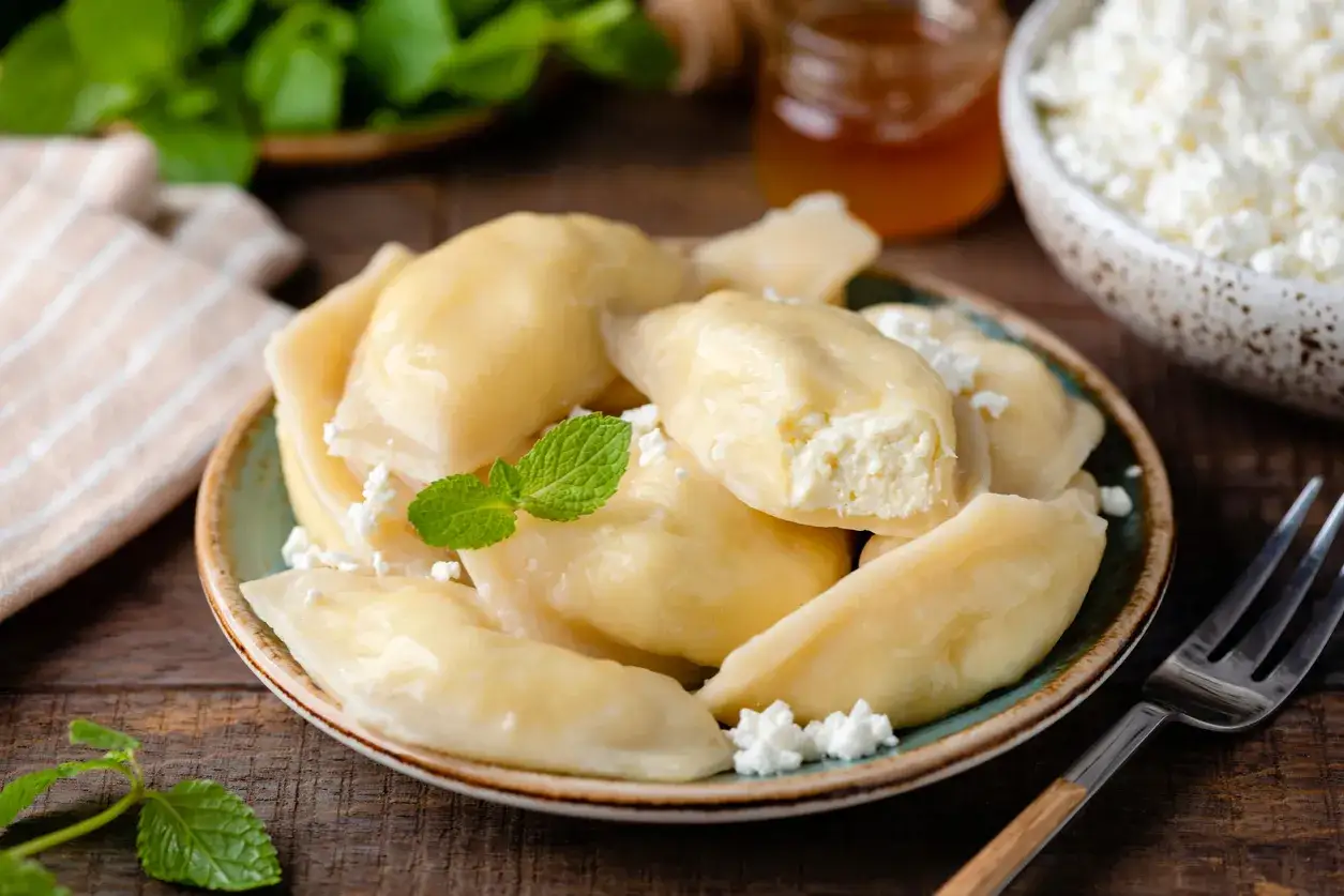 Talerz pełen pysznych, domowych pierogów z serem, udekorowany miętą. Obok miseczka z twarogiem i słoiczek miodu.