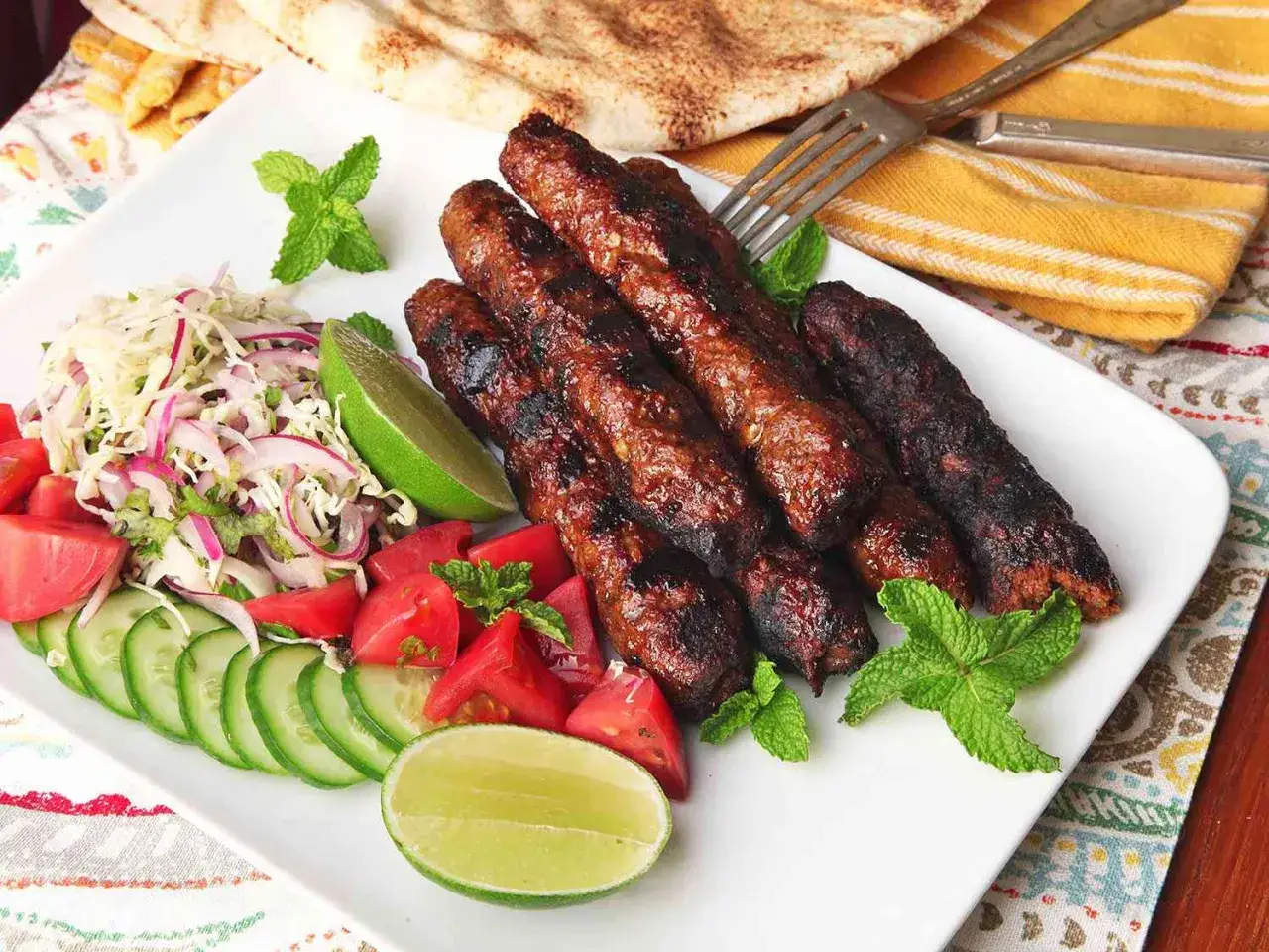 Grillowany kebab z mięsa mielonego, podany z sałatką z kapusty, pomidorów, ogórków, limonki i listkami mięty. Obok pieczywo pita.