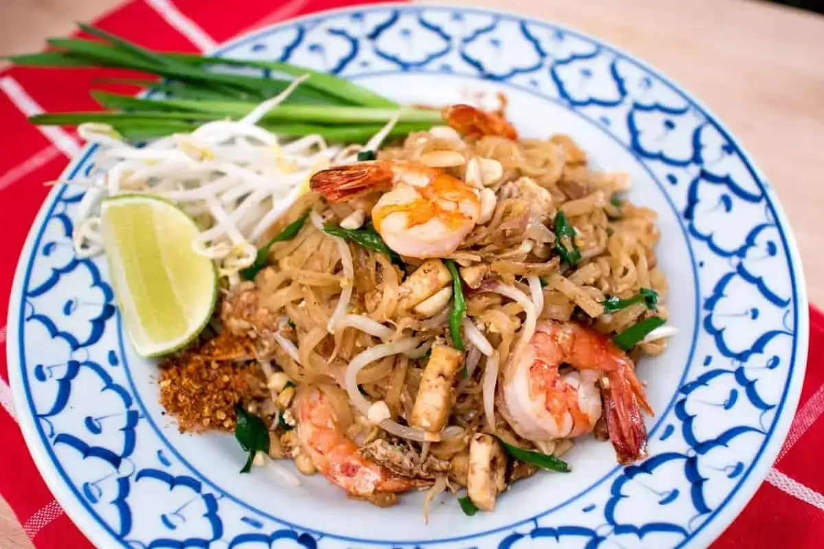 Pyszny pad thai przepis z krewetkami, kiełkami fasoli mung, szczypiorkiem i ćwiartką limonki na talerzu z niebieskim wzorem.