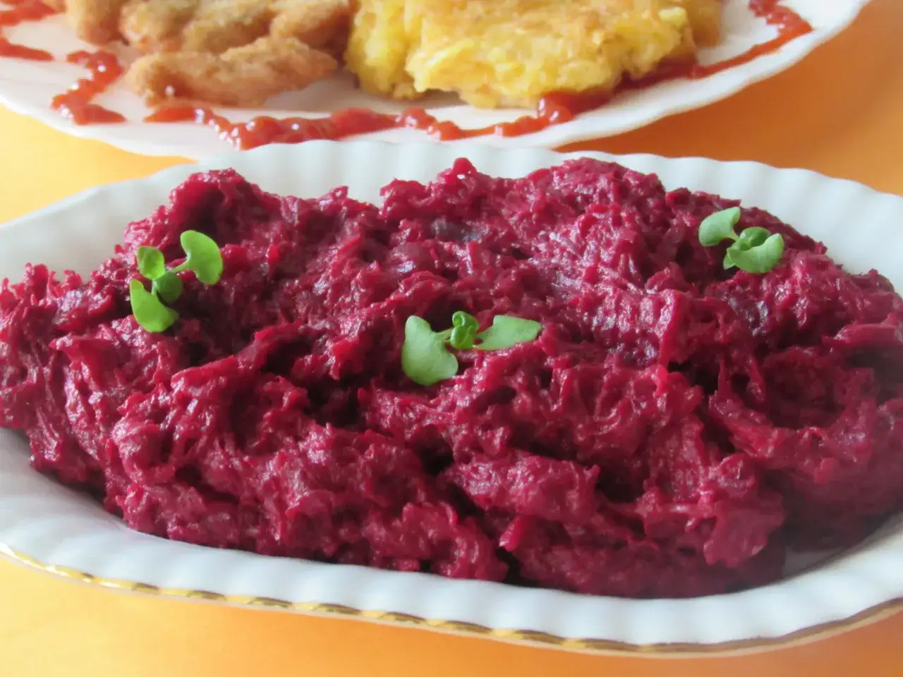 Pyszne, purpurowe buraczki na ciepło, udekorowane listkami zi&oacute;ł. W tle widać kotlety i placki ziemniaczane.