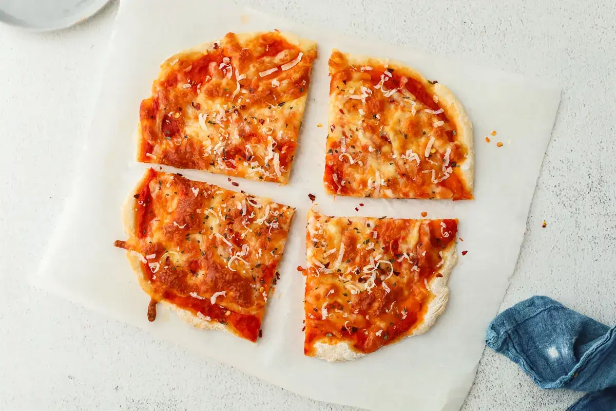 Cztery kawałki pizzy z serka wiejskiego, posypane startym serem i ziołami, leżą na papierze do pieczenia.
