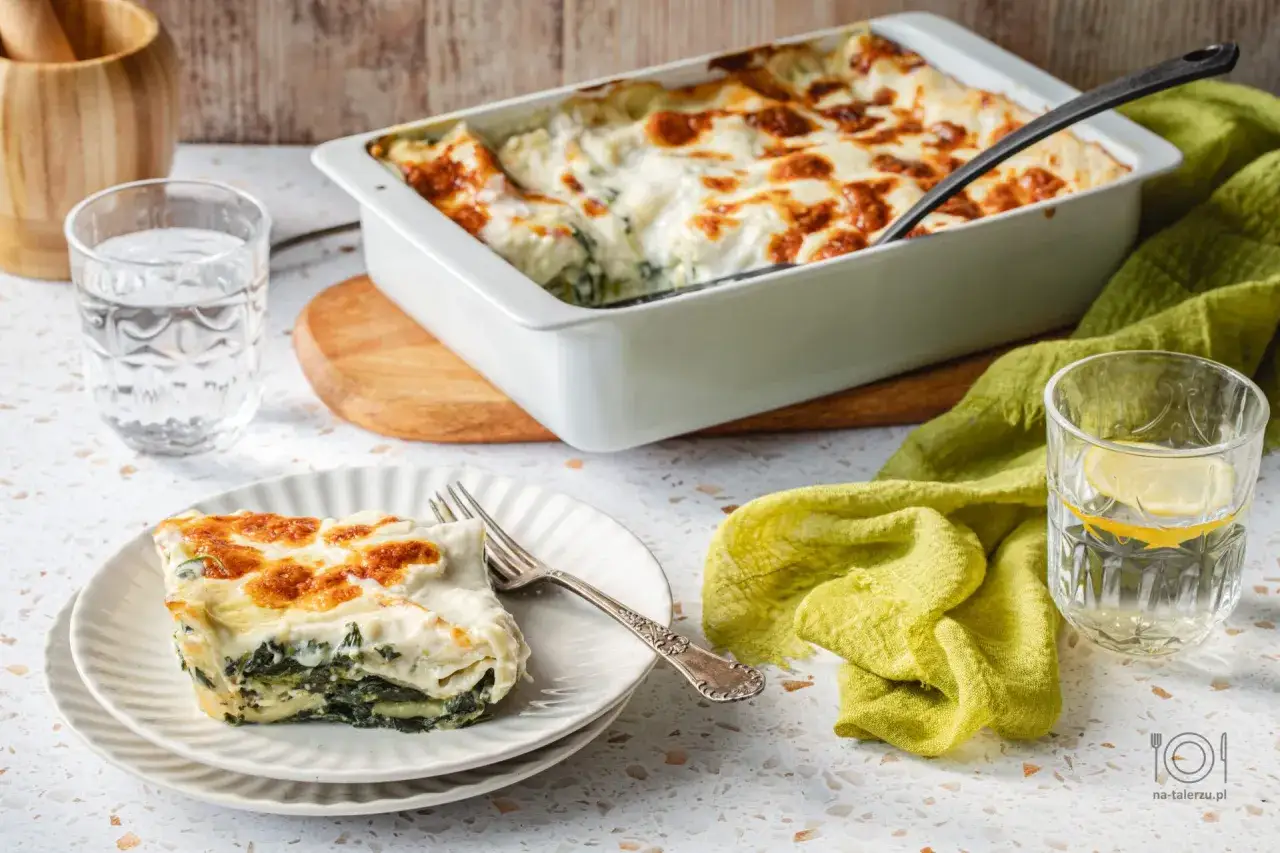 Pyszne lasagne ze szpinakiem, zapieczone na złoto, podane na talerzu obok szklanki wody z cytryną.