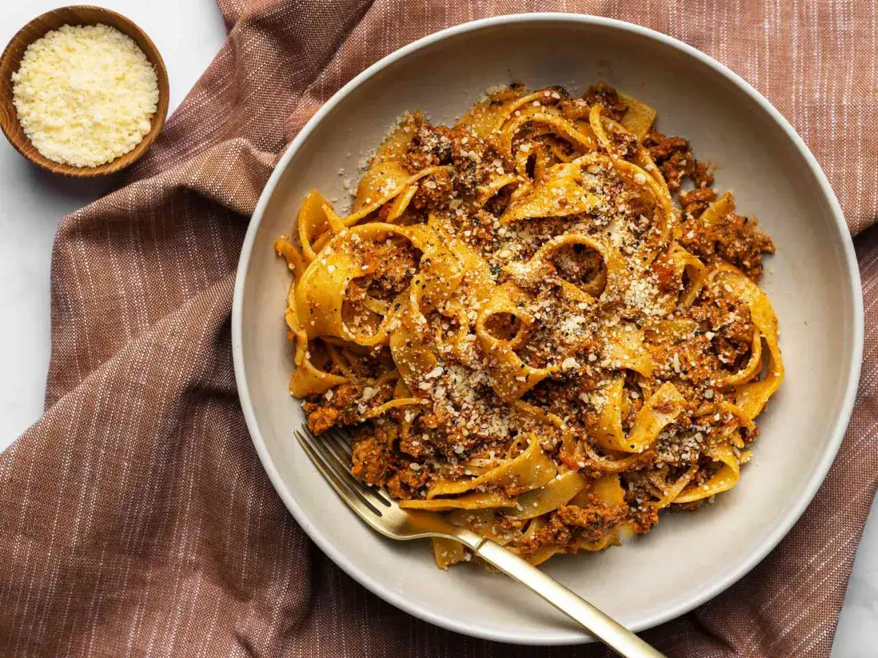Talerz tagliatelle z bogatym, mięsnym sosem do makaronu, posypany parmezanem. Obok miseczka z tartym serem.