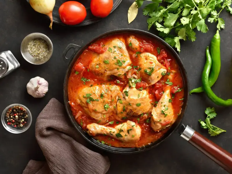 Pyszny kurczak w sosie pomidorowym, duszony z cebulą i przyprawami, udekorowany natką pietruszki. Obok zielona papryczka chili i pomidory.